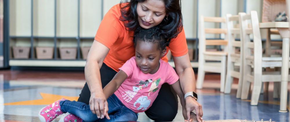Imagine Early Education teacher kneeling beside a toddler on the classroom floor, guiding her hand as she strums an electric guitar during music class.