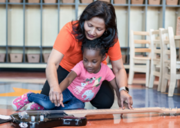 Imagine Early Education teacher kneeling beside a toddler on the classroom floor, guiding her hand as she strums an electric guitar during music class.
