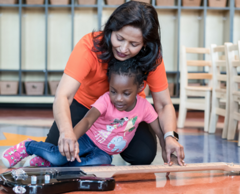 Imagine Early Education teacher kneeling beside a toddler on the classroom floor, guiding her hand as she strums an electric guitar during music class.