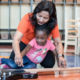 Imagine Early Education teacher kneeling beside a toddler on the classroom floor, guiding her hand as she strums an electric guitar during music class.
