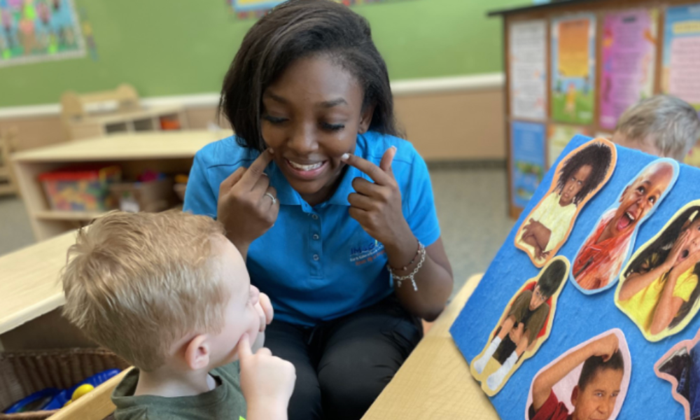 Imagine Early Education teacher smiling and pointing to her cheeks as a young boy copies her, with a felt board of children’s faces beside them in a preschool classroom.