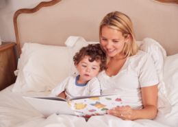 Smiling mother and curly-haired toddler in white shirts reading a colorful picture book together while sitting up in bed against white pillows