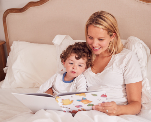 Smiling mother and curly-haired toddler in white shirts reading a colorful picture book together while sitting up in bed against white pillows