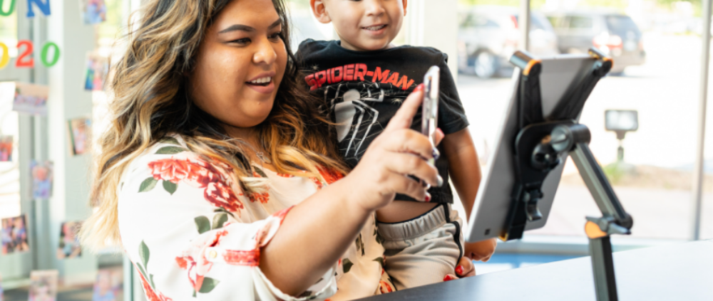 Smiling parent holding her young son on her hip as she uses her phone at a mounted tablet check-in station inside an Imagine Early Education center.
