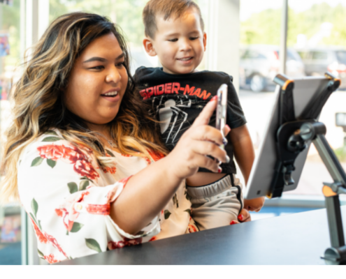 Smiling parent holding her young son on her hip as she uses her phone at a mounted tablet check-in station inside an Imagine Early Education center.