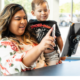 Smiling parent holding her young son on her hip as she uses her phone at a mounted tablet check-in station inside an Imagine Early Education center.