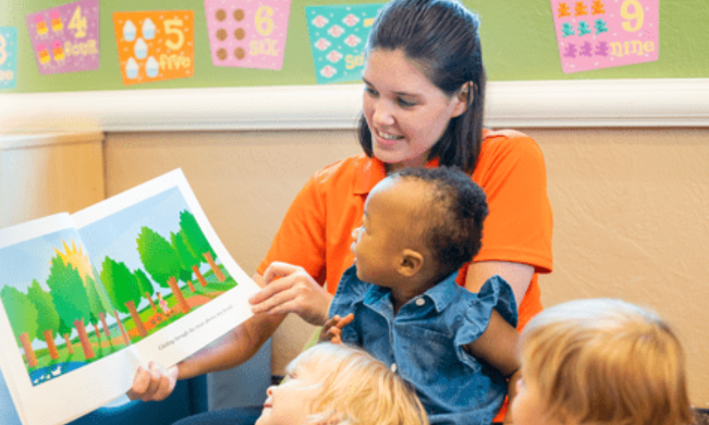 Imagine Early Education teacher holding a toddler on her lap while reading a picture book aloud to a small group of children in a classroom decorated with number posters.